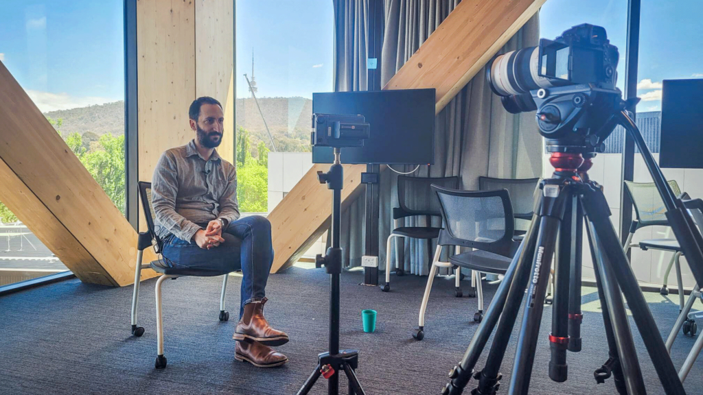 A man is sitting on a chair, there is a camera on a tripod and lighting on stands.