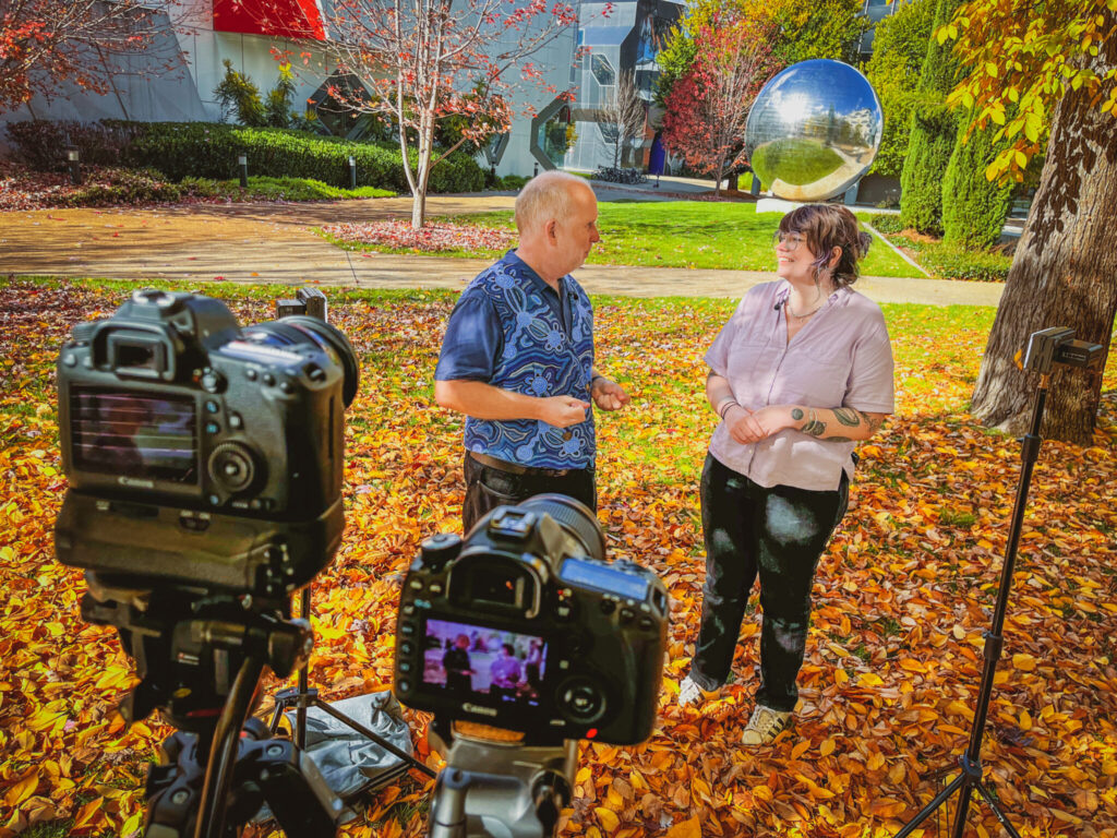 A man and a woman stand facing each other in an outside location, the ground is covered with brightly coloured autumn leaves, two cameras are visible in the foreground, mounted on tripods.