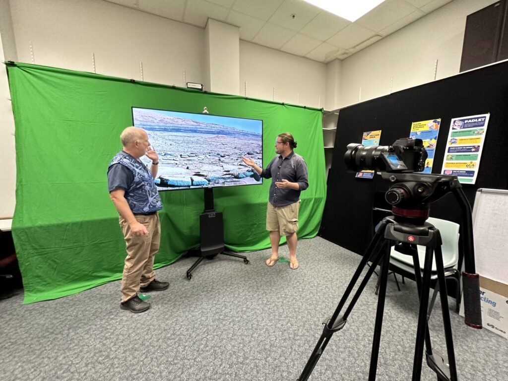 Two men stand in front of a large television screen, a green backdrop is behind them, a video camera is on a stand in front of them. 