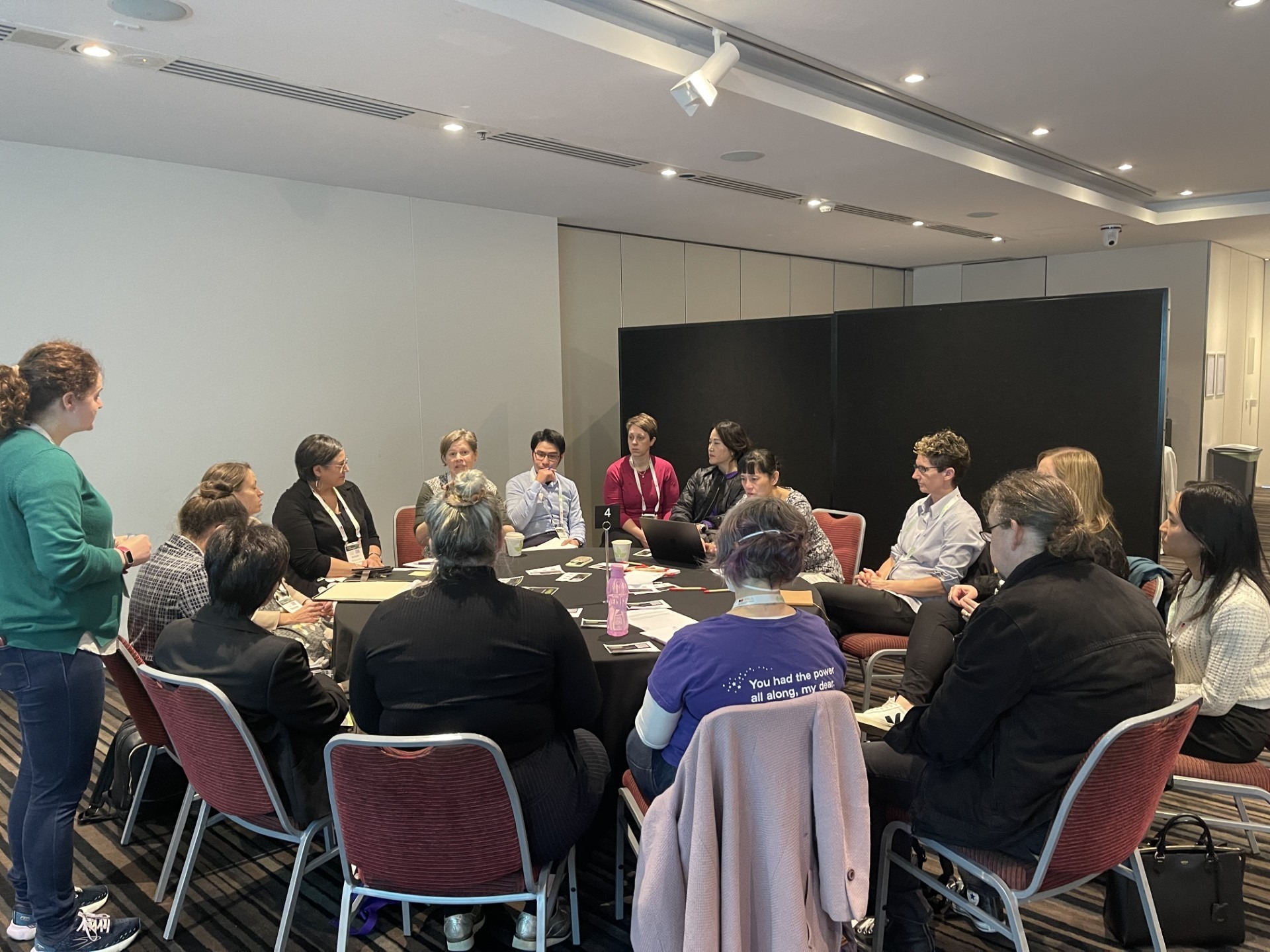 Sue Sharpe sits at a large round table, speaking with 14 participants, while her co-presenter Mel Saligari stands looks on.