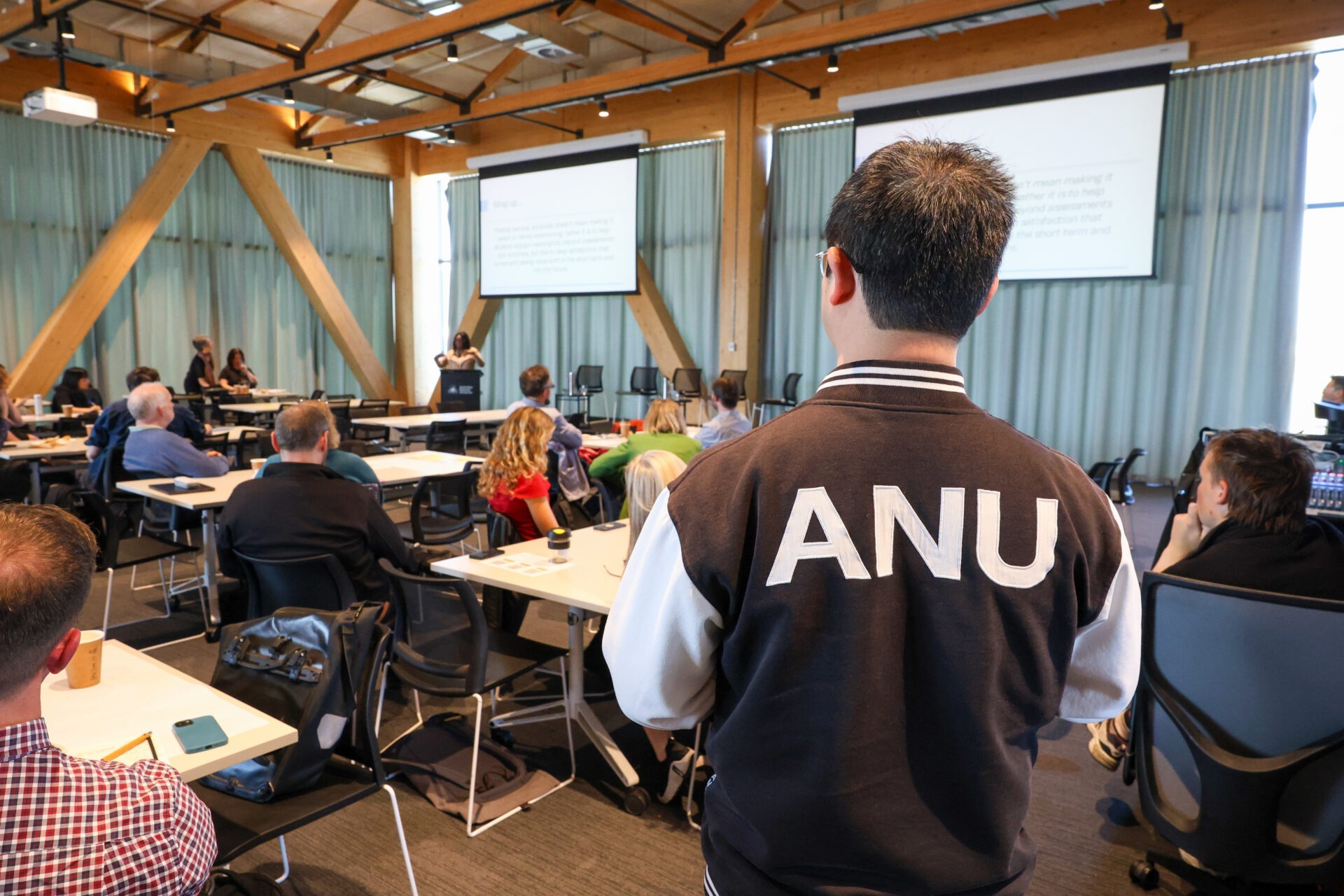 An attendee with ANU lettering on their jacket looks towards the presentation area.